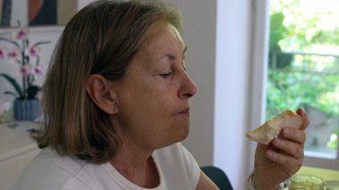 Yaşlı kadın, mutfak masasında otururken bir ısırık almayı seviyor. Sabah ışığı pencereden içeri giriyor ve arka plandaki rafta orkide var.