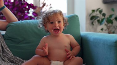 Yeni yürümeye başlayan çocuk keyifle el çırpıyor samimi oturma odasındaki deniz mavisi kanepede otururken, bitkilerle çevrili ve neşeli anlarda yumuşak doğal ışıkla.