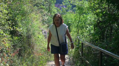 Yaşlı kadın orman yolunda tek başına yürüyor, yemyeşil ile çevrili dar bir yolda yürüyor ve güneşli yaz gününde elinde küçük bir çapraz ceset torbası tutuyor.