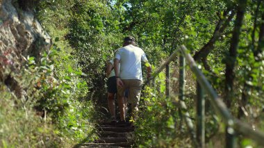 Yaşlı çift yaz yürüyüşü sırasında sık bitki örtüsü, ağaçlar ve korkuluklarla çevrili kırsal taş basamaklarda tırmanıyor.