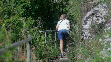 Vahşi bitkiler ve kayalık yamaçlarla çevrili orman yolunda yürüyen bir kadının arka görüntüsü yaz boyunca metal korkuluklarla dar patikalara tırmanıyor.
