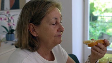 Yaşlı kadın, mutfak masasında otururken bir ısırık almayı seviyor. Sabah ışığı pencereden içeri giriyor ve arka plandaki rafta orkide var.