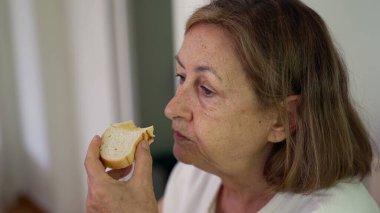 Beyaz duvar arkasında otururken sandviçi ısıran, tarafsız tişört giyen ve yemeğine odaklanmış yaşlı bir kadının yakın çekimi.