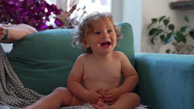 Neşeli çocuk, başı öne eğik gülerken kapalı alandaki deniz mavisi koltukta, bitkilerle çevrili ve rahat aile ortamında yumuşak doğal ışık altında.