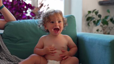 Yeni yürümeye başlayan çocuk keyifle el çırpıyor samimi oturma odasındaki deniz mavisi kanepede otururken, bitkilerle çevrili ve neşeli anlarda yumuşak doğal ışıkla.