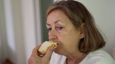 Beyaz duvar arkasında otururken sandviçi ısıran, tarafsız tişört giyen ve yemeğine odaklanmış yaşlı bir kadının yakın çekimi.