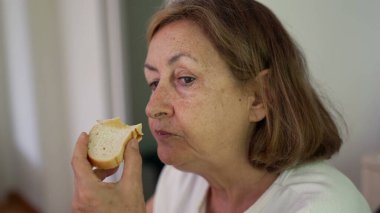 Beyaz duvar arkasında otururken sandviçi ısıran, tarafsız tişört giyen ve yemeğine odaklanmış yaşlı bir kadının yakın çekimi.