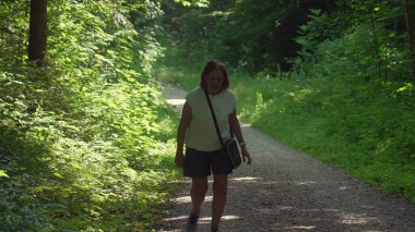 Yaşlı bir kadın, yemyeşil ormandaki çakıllı patikada tek başına yürüyor güneşli yaz sabahında, ağaçlar ve bitkilerle çevrili, huzur içinde tek başına yürüyüş yapıyor.