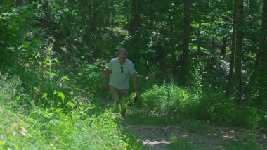 Yaşlı adam ormanda yokuş yukarı yürüyor, yeşil yapraklarla çevrili toprak yolda odaklanmış bir ifadeyle yürüyor doğadaki tek başına yaz fitness aktivitesi sırasında.