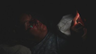 Yaşlı kadın gece yatağında akıllı telefon kullanarak uzanıyor, yüzü karanlık yatak odasındaki ekran ışığıyla aydınlanıyor, gözlük takıyor ve cihaza odaklanıyor.