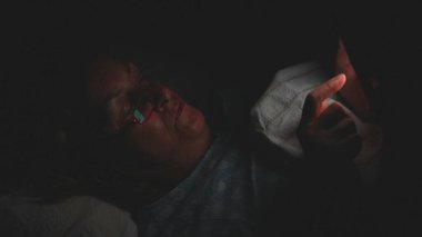 Yaşlı kadın gece yatağında akıllı telefon kullanarak uzanıyor, yüzü karanlık yatak odasındaki ekran ışığıyla aydınlanıyor, gözlük takıyor ve cihaza odaklanıyor.