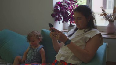 Anne, bebek koltuğunda otururken akıllı telefon kullanıyor. Oturma odasında doğal ışık, bitkiler ve günlük aile etkileşimi olan sakin bir ev kadını.