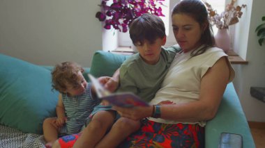 Anne, kanepede iki küçük çocukla kitap okuyor, daha yeni yürümeye başlayan bebek, büyük çocuk onu takip ederken, sıcak oturma odasında aile bağı kurma zamanı.