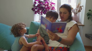Anne, kanepede iki küçük çocukla kitap okuyor, daha yeni yürümeye başlayan bebek, büyük çocuk onu takip ederken, sıcak oturma odasında aile bağı kurma zamanı.