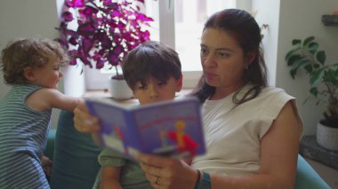 Anne, çocuklarıyla oturma odasında resim kitabı okuyor, küçük çocuk seyrediyor ve gülümsüyor, büyük çocuk ise sayfalara konsantre oluyor, sıcak eğitim anı.