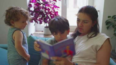 Anne, çocuklarıyla oturma odasında resim kitabı okuyor, küçük çocuk seyrediyor ve gülümsüyor, büyük çocuk ise sayfalara konsantre oluyor, sıcak eğitim anı.