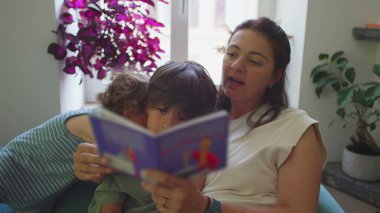 Anne, çocuklarıyla oturma odasında resim kitabı okuyor, küçük çocuk seyrediyor ve gülümsüyor, büyük çocuk ise sayfalara konsantre oluyor, sıcak eğitim anı.