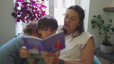 Anne, çocuklarıyla oturma odasında resim kitabı okuyor, küçük çocuk seyrediyor ve gülümsüyor, büyük çocuk ise sayfalara konsantre oluyor, sıcak eğitim anı.
