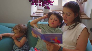 Anne, oturma odasındaki kanepede oturan iki çocuğa masal kitabı okuyor. Çocuğun dikkati dağılıyor. Büyük çocuk eğiliyor.
