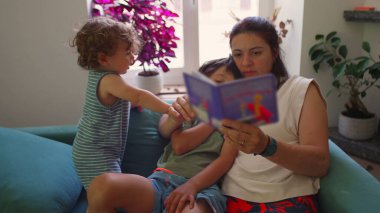 Anne kanepede iki çocukla oturup resim kitabı okurken, bir çocuk kollarına sarılıp dikkatle dinlerken, diğeri de sıcak bir aile salonunda onların yanında şekerleme yapıyor.