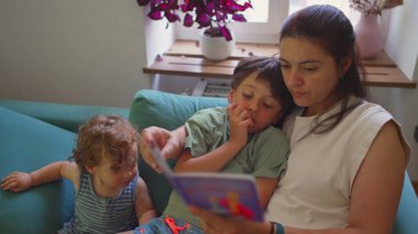 Anne, oturma odasındaki kanepede oturan iki çocuğa masal kitabı okuyor. Çocuğun dikkati dağılıyor. Büyük çocuk eğiliyor.