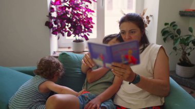 Anne kanepede iki çocukla oturup resim kitabı okurken, bir çocuk kollarına sarılıp dikkatle dinlerken, diğeri de sıcak bir aile salonunda onların yanında şekerleme yapıyor.