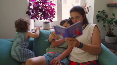 Anne kanepede iki çocukla oturup resim kitabı okurken, bir çocuk kollarına sarılıp dikkatle dinlerken, diğeri de sıcak bir aile salonunda onların yanında şekerleme yapıyor.