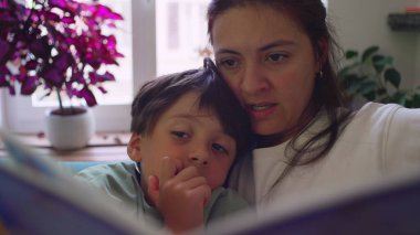 Anne, küçük oğluna kanepede sarılırken pencere kenarında kitap okuyor. Her ikisi de oturma odasında doğal ışık ve saksı bitkileriyle dolu resimli kitaplara odaklanmış.