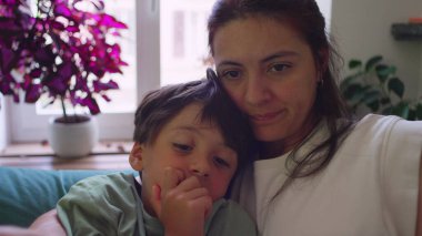 Anne, küçük oğluna kanepede sarılırken pencere kenarında kitap okuyor. Her ikisi de oturma odasında doğal ışık ve saksı bitkileriyle dolu resimli kitaplara odaklanmış.