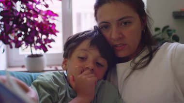 Anne, küçük oğluna kanepede sarılırken pencere kenarında kitap okuyor. Her ikisi de oturma odasında doğal ışık ve saksı bitkileriyle dolu resimli kitaplara odaklanmış.