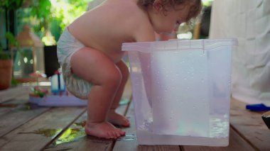 Bebek çıplak ayakla ayakta duruyor, temiz plastik bir çöp kutusunu tutuyor, yemyeşil bahçe ortamında dağınık su oyuncaklarıyla ıslak ahşap verandada oynuyor.