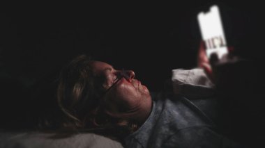 Yaşlı kadın karanlıkta yatakta uzanmış, ışığı yansıtan gözlüklerle gözünün önünde telefonu tutarken parlayan akıllı telefon ekranına bakıyor.