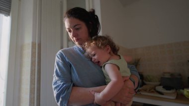 Anne, yumuşak pencere ışığı ile sıcak mutfakta kalçasında kıvırcık saçlı bebek kucağında, her ikisi de nazik bir ifadeyle tezgaha doğru bakıyor