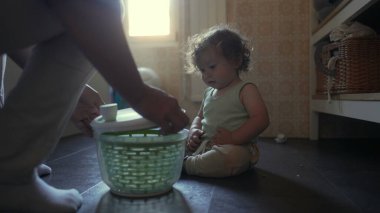 Küçük çocuk mutfak zemininde oturmuş salata çeviricisini seyrederken yetişkin asistanları sıcak ev ortamında