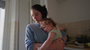 Anne, yumuşak pencere ışığı ile sıcak mutfakta kalçasında kıvırcık saçlı bebek kucağında, her ikisi de nazik bir ifadeyle tezgaha doğru bakıyor