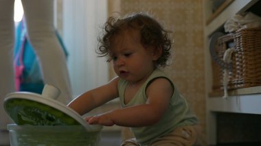 Kıvırcık saçlı çocuk mutfakta salata fırıldağıyla dikkatle oynuyor. Erken gelişim aşamasında yerde otururken sapını çevirmeye odaklanmış.