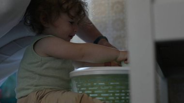 Kıvırcık saçlı çocuk, yetişkin elinin yardımıyla salata fırıldağıyla oynarken, oyun zamanındaki ilk keşifler sırasında etkileşim ve destek anı