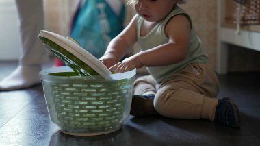 Kıvırcık saçlı meraklı çocuk, mutfak zemininde salata fırıldağıyla oynuyor, erken çocukluk gelişimi sırasında oturmuş ve araştırma yapıyor.