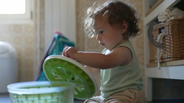 Kıvırcık saçlı çocuk mutfakta salata fırıldağıyla dikkatle oynuyor. Erken gelişim aşamasında yerde otururken sapını çevirmeye odaklanmış.
