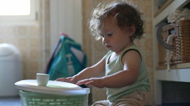 Kıvırcık saçlı çocuk mutfakta salata fırıldağıyla dikkatle oynuyor. Erken gelişim aşamasında yerde otururken sapını çevirmeye odaklanmış.