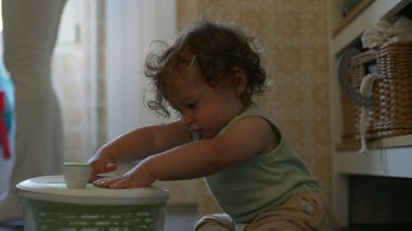 Kıvırcık saçlı çocuk mutfakta salata fırıldağıyla dikkatle oynuyor. Erken gelişim aşamasında yerde otururken sapını çevirmeye odaklanmış.