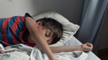 Yatakta uzandıktan sonra kollarını açarak uzanan genç çocuk, uykulu bir ifadeyle ve yumuşak yatak odası ışığında kıvırcık saçlarıyla yavaşça uyanıyor.