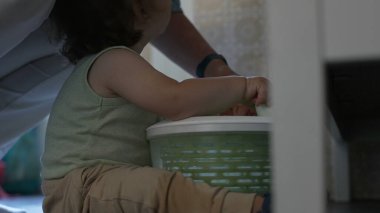 Kıvırcık saçlı çocuk, yetişkin elinin yardımıyla salata fırıldağıyla oynarken, oyun zamanındaki ilk keşifler sırasında etkileşim ve destek anı