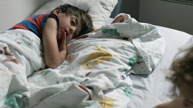 Kıvırcık saçlı çocuk, yatağın yanında durup aşağı bakarken, büyük kardeş battaniyenin altından izliyor, kardeş arasında geçen sakin bir sabah anı.