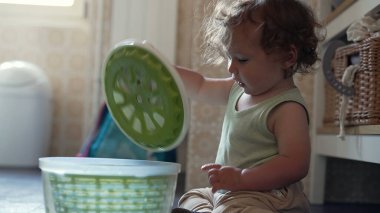 Kıvırcık saçlı meraklı çocuk, mutfak zemininde salata fırıldağıyla oynuyor, erken çocukluk gelişimi sırasında oturmuş ve araştırma yapıyor.