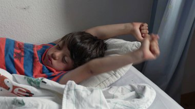 Yatakta uzandıktan sonra kollarını açarak uzanan genç çocuk, uykulu bir ifadeyle ve yumuşak yatak odası ışığında kıvırcık saçlarıyla yavaşça uyanıyor.