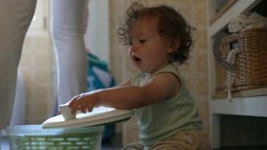 Kıvırcık saçlı çocuk mutfakta salata fırıldağıyla dikkatle oynuyor. Erken gelişim aşamasında yerde otururken sapını çevirmeye odaklanmış.