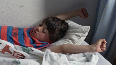 Yatakta uzandıktan sonra kollarını açarak uzanan genç çocuk, uykulu bir ifadeyle ve yumuşak yatak odası ışığında kıvırcık saçlarıyla yavaşça uyanıyor.