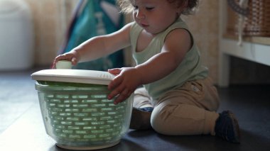 Kıvırcık saçlı meraklı çocuk, mutfak zemininde salata fırıldağıyla oynuyor, erken çocukluk gelişimi sırasında oturmuş ve araştırma yapıyor.