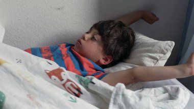 Yatakta uzandıktan sonra kollarını açarak uzanan genç çocuk, uykulu bir ifadeyle ve yumuşak yatak odası ışığında kıvırcık saçlarıyla yavaşça uyanıyor.